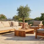 A luxurious wooden outdoor lounge set with neutral cushions on a sun-drenched stone patio, surrounded by lush garden and flowers.