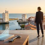 Une femme d'affaires observe La Rochelle. Au premier plan, un smartphone affiche un graphique de croissance et une clé, sous un soleil couchant.