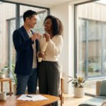 Couple mixte souriant tenant des clés dans un salon lumineux et moderne. Des documents sont sur la table basse.