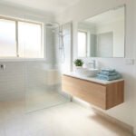 Salle de bain moderne et lumineuse avec douche à l'italienne, meuble vasque en bois clair, miroir et femme souriante.