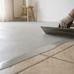Un professionnel applique un enduit de ragréage gris sur un carrelage existant avec une lisseuse pour le rénover.