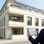Homme en costume regardant un immeuble moderne avec panneaux solaires, tablette affichant plans.