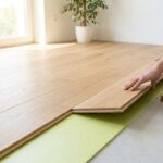 Gros plan sur l'installation de parquet flottant clair, avec des mains alignant une lame sur une sous-couche verte. Outils à droite.