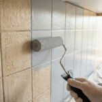 Personne peignant des carreaux de faïence beige avec un rouleau et de la peinture grise pour rénover une cuisine.