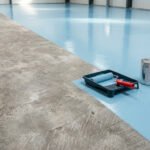 Sol en béton partiellement peint en bleu clair brillant. Un rouleau et un pot de peinture sont visibles, illustrant l'application d'une peinture de protection.