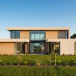 Façade d'une maison moderne avec bardage bois et grandes baies vitrées, jardin verdoyant et mer à l'horizon sous ciel bleu.