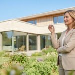 Une femme élégante souriante devant une maison moderne, tenant les clés de la propriété dans sa main.