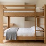 Lit superposé adulte en bois clair, style moderne et épuré. Draps blancs, plaid gris. Bureau intégré et lumière naturelle.